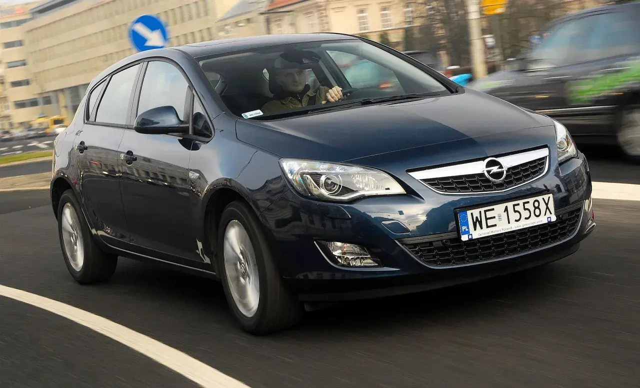Ciemnoniebieski Opel Astra J 1.6 benzyna w ruchu miejskim. Kierowca w czapce prowadzi auto.