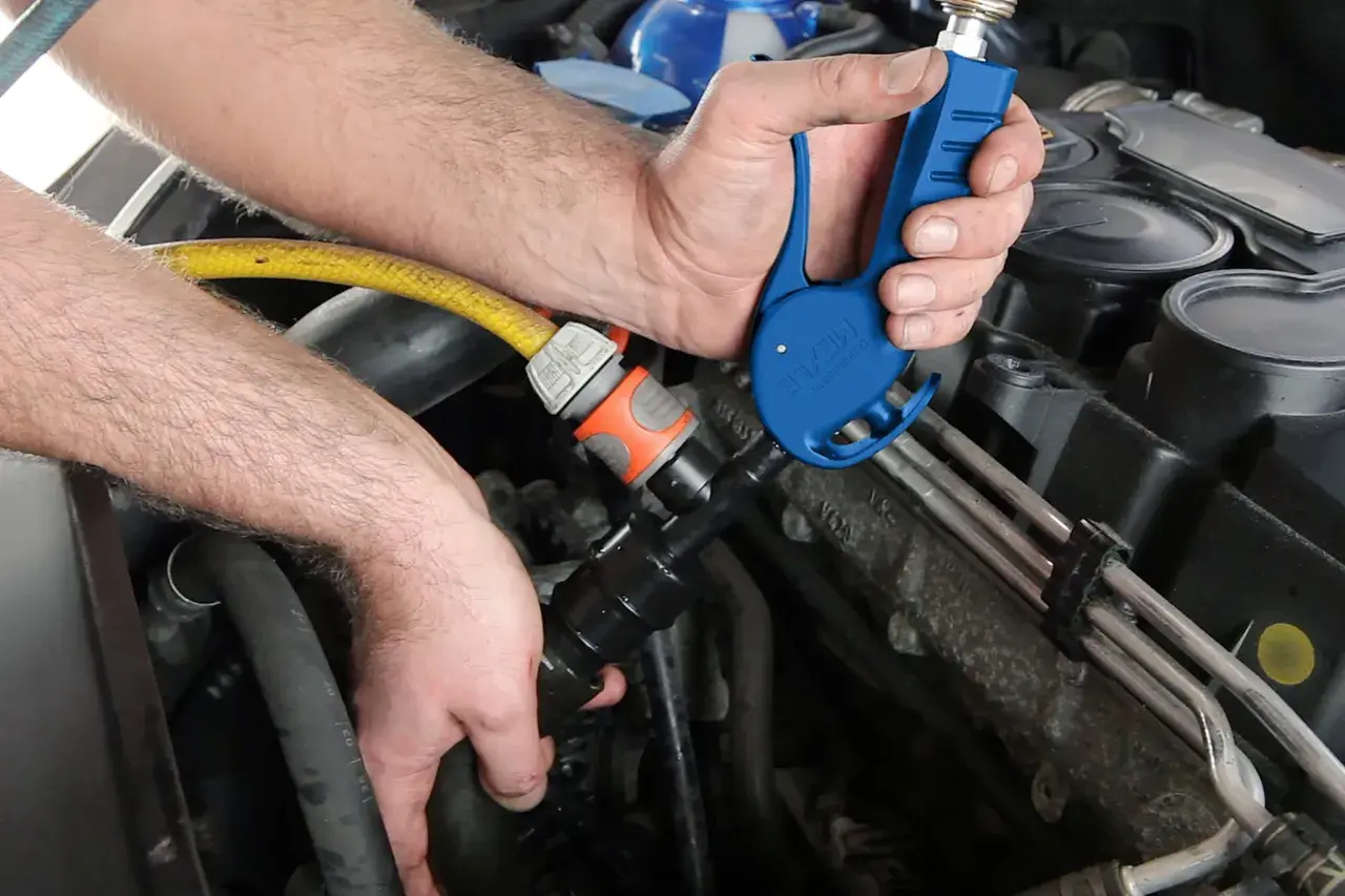 Ręce mechanika podłączają niebieskie narzędzie do węża, przygotowując środek do płukania układu chłodzenia.