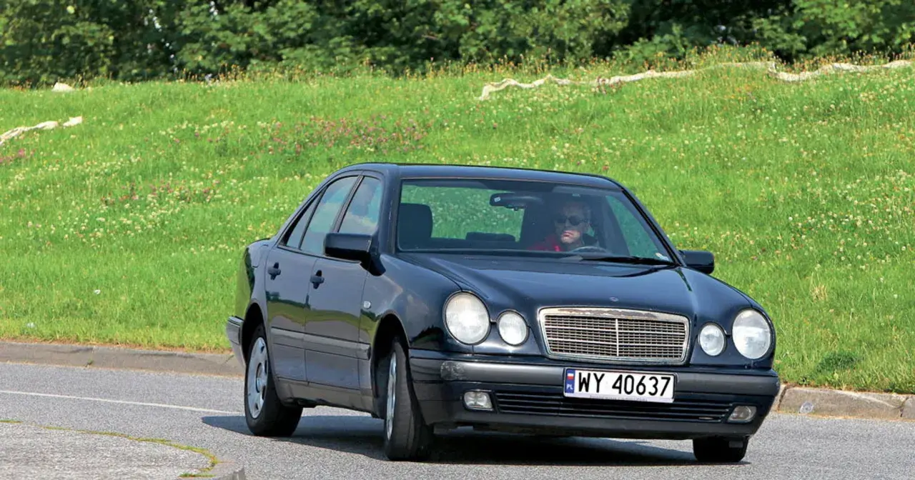 Czarny Mercedes W210 z silnikiem om606 jedzie po drodze, za nim zielone, kwieciste wzgórze.
