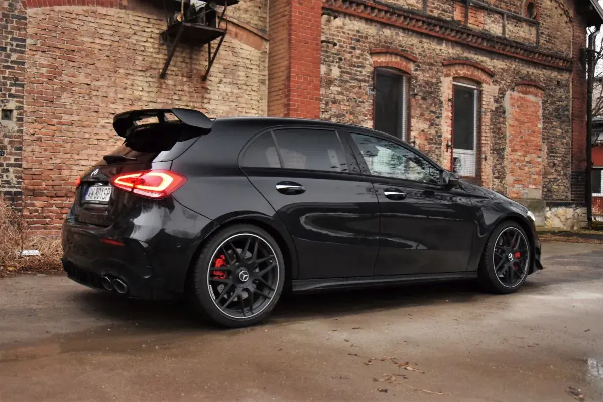 Czarny Mercedes-AMG A45s z dużym spojlerem i czerwonymi zaciskami hamulcowymi stoi przed starą ceglaną kamienicą.