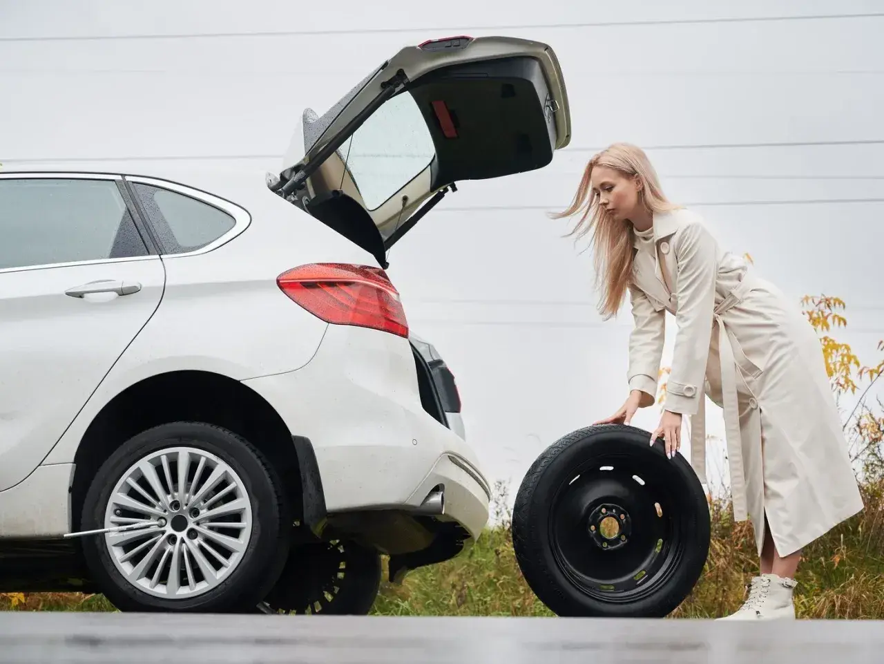 Kobieta wyjmuje koło zapasowe z bagażnika białego auta. W tle widać drogę i jesienne drzewa. To może być pomocne, jak zdjąć koło zapasowe Citroen Jumper.