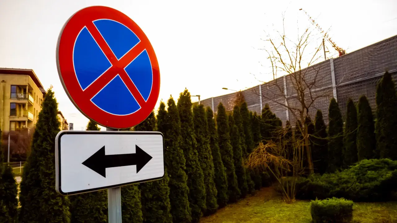 Znak zakaz zatrzymywania z tabliczką wskazującą kierunki. W tle budynek i żywopłot.