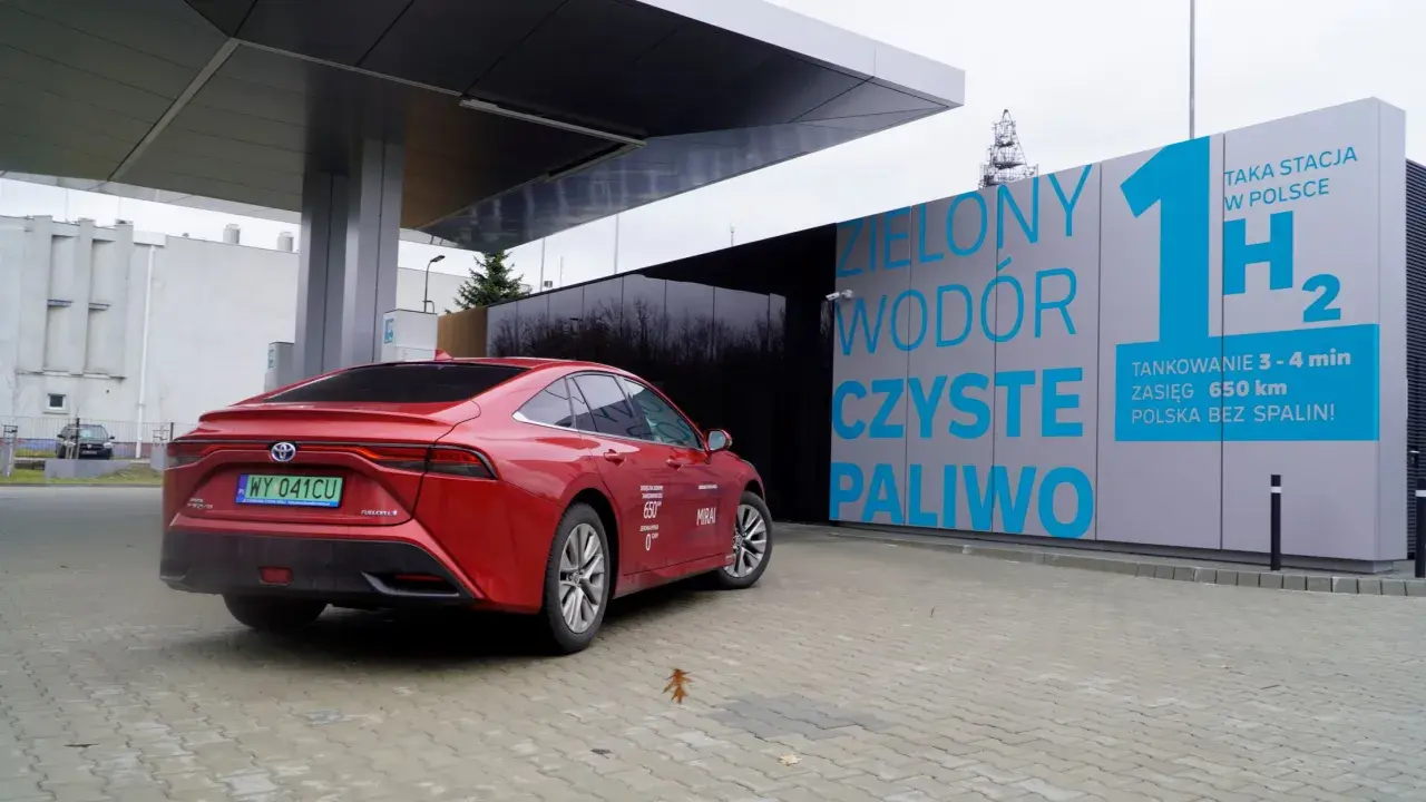 Czerwona Toyota Mirai na stacji tankowania wodoru. "Zielony wodór, czyste paliwo" - hasła na budynku stacji.