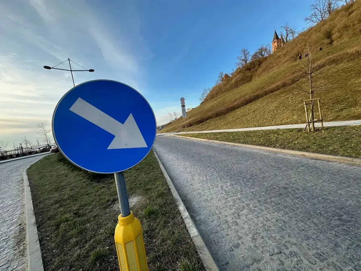 Niebieski znak drogowy ze strzałką w prawo na tle zielonego wzgórza i nieba.