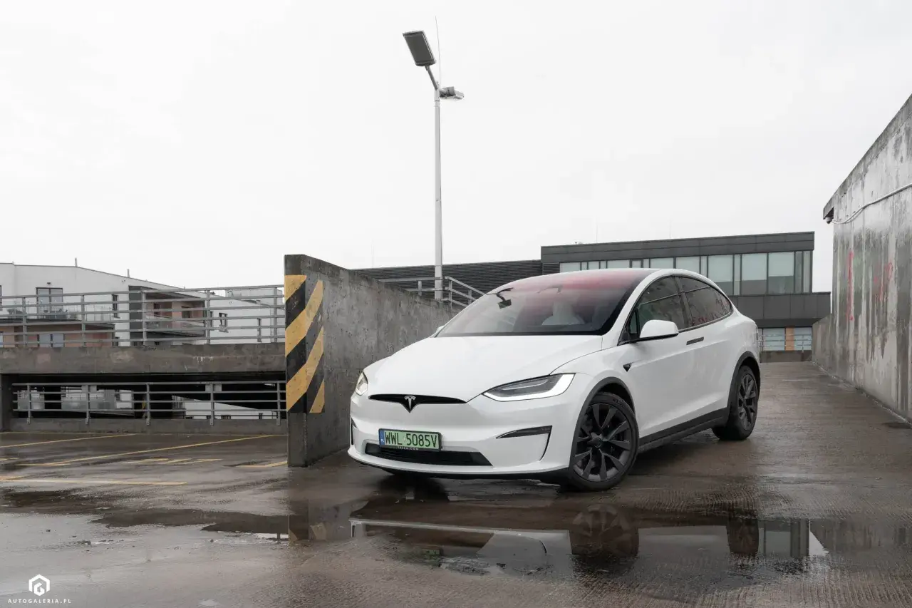 Biały Tesla Model X na mokrym parkingu, z widokiem na budynki i latarnię.