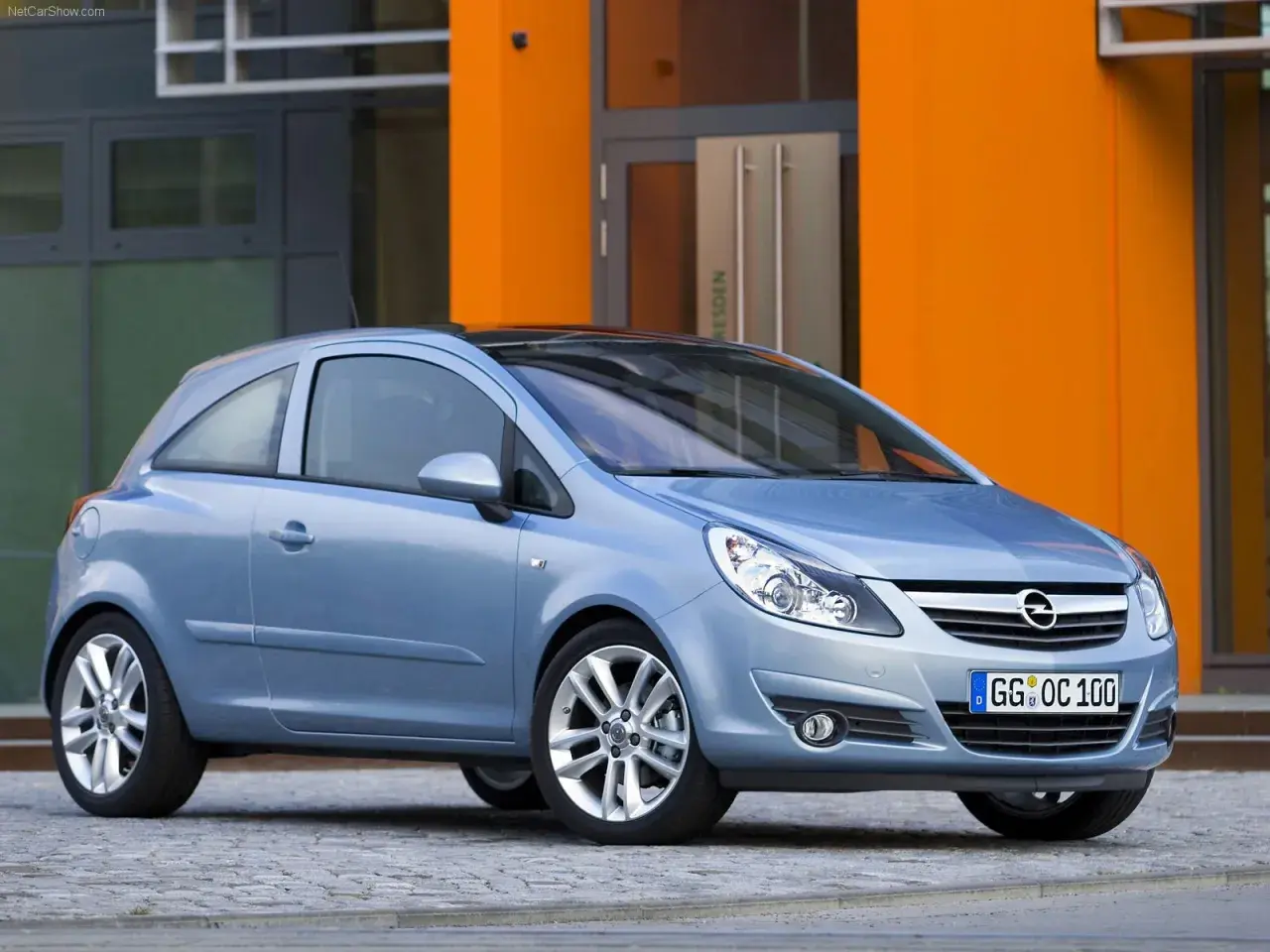 Niebieski Opel Corsa D 1.2 benzyna stoi przed pomarańczowym budynkiem.