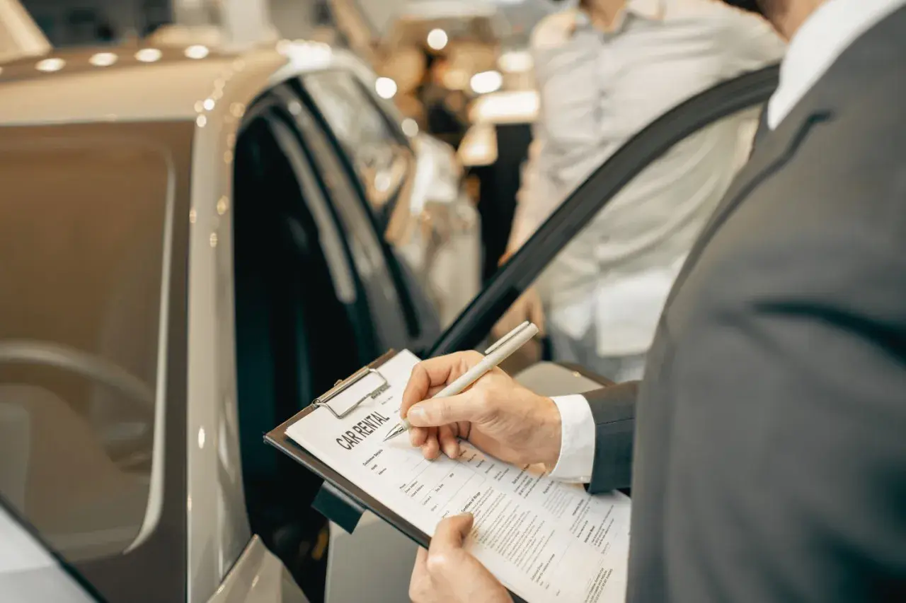 Mężczyzna w garniturze podpisuje dokument "CAR RENTAL" przy luksusowym samochodzie.