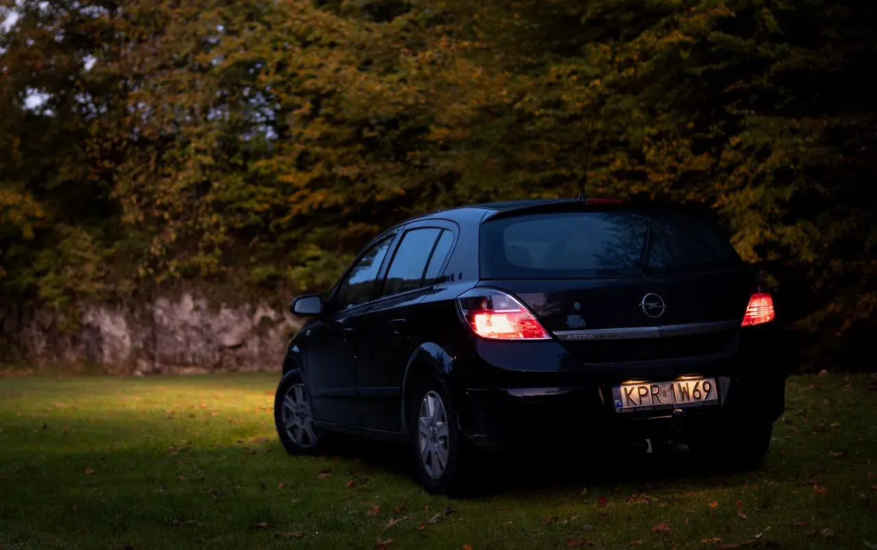 Czarny Opel Astra H 1.6 benzyna zaparkowany na trawie, w tle jesienne drzewa.