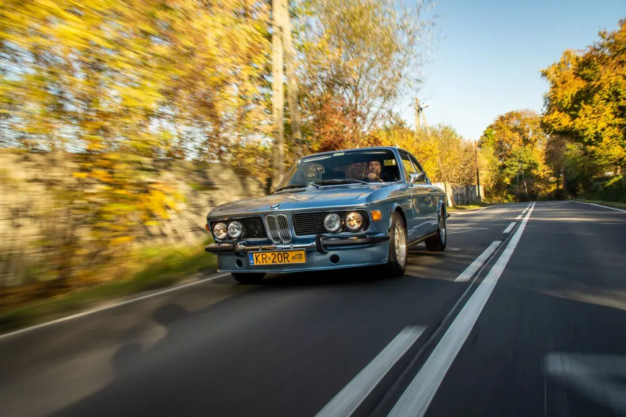 Błękitne BMW E9 z kierowcą jedzie jesienną drogą, otoczone kolorowymi drzewami.