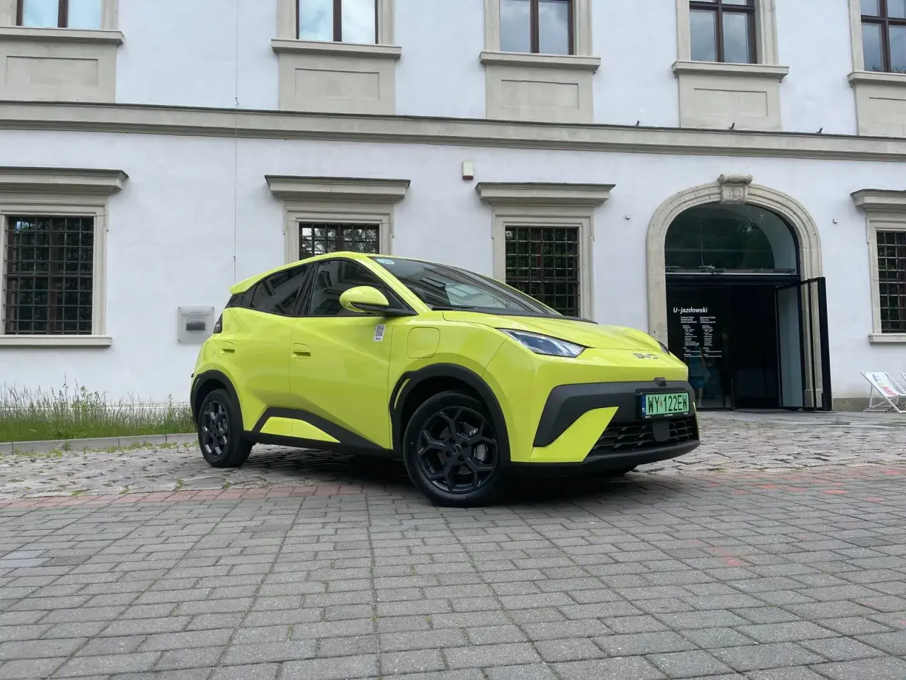 Jaskrawożółty BYD Seagull zaparkowany na bruku przed budynkiem z łukowym wejściem.