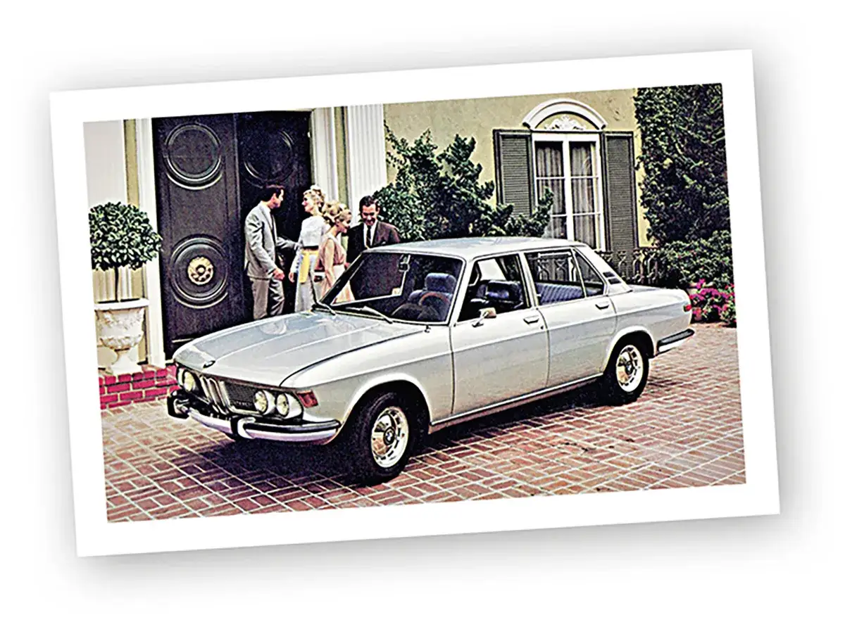 Elegancki, srebrny sedan BMW E9 z lat 70. stoi przed domem, a obok niego ludzie w strojach z epoki.