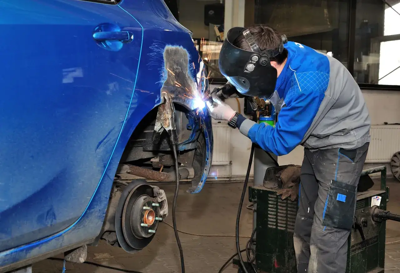Mechanik w masce spawa błotnik, pokazując, jak załatać dziury w samochodzie. Iskry sypią się podczas naprawy.