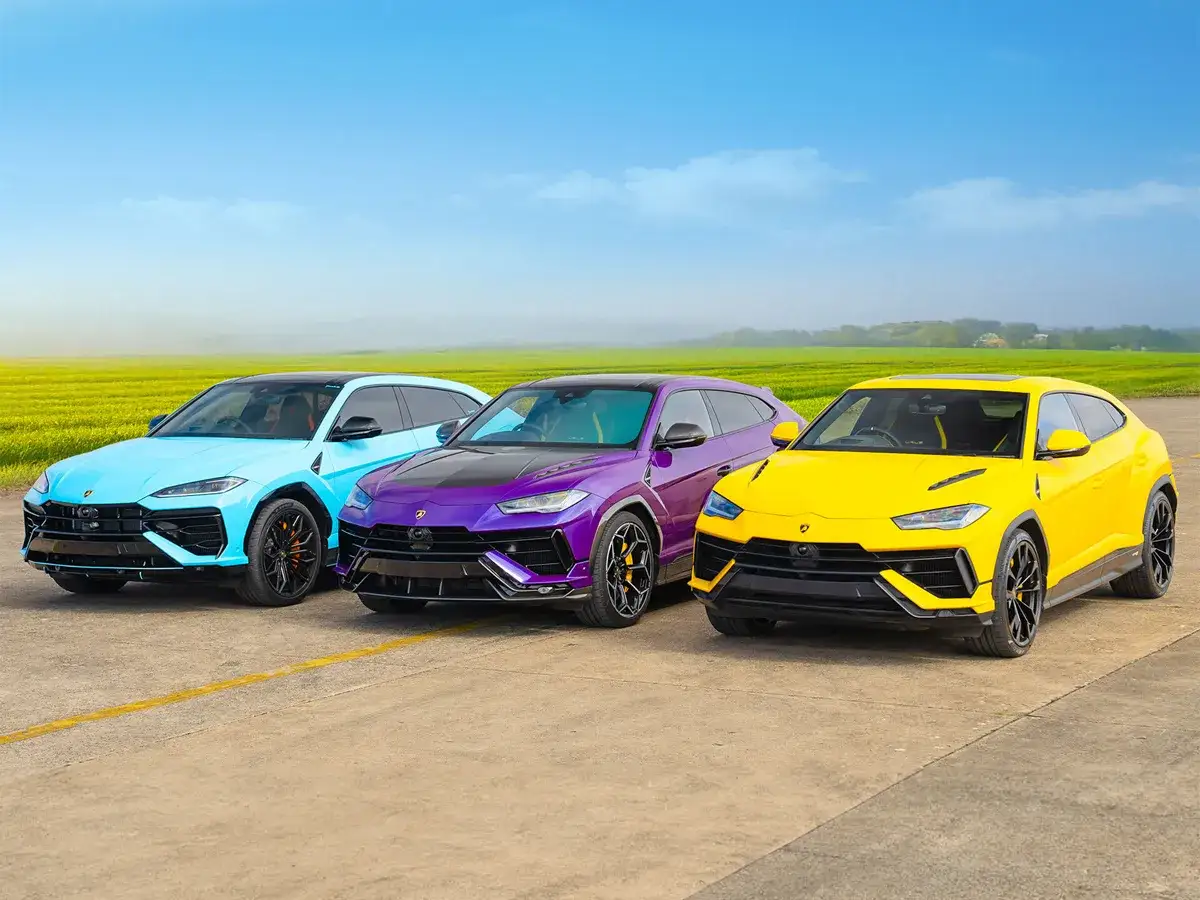 Trzy Lamborghini Urus w jaskrawych kolorach: błękitnym, fioletowym i żółtym, stoją obok siebie na tle zielonych pól.