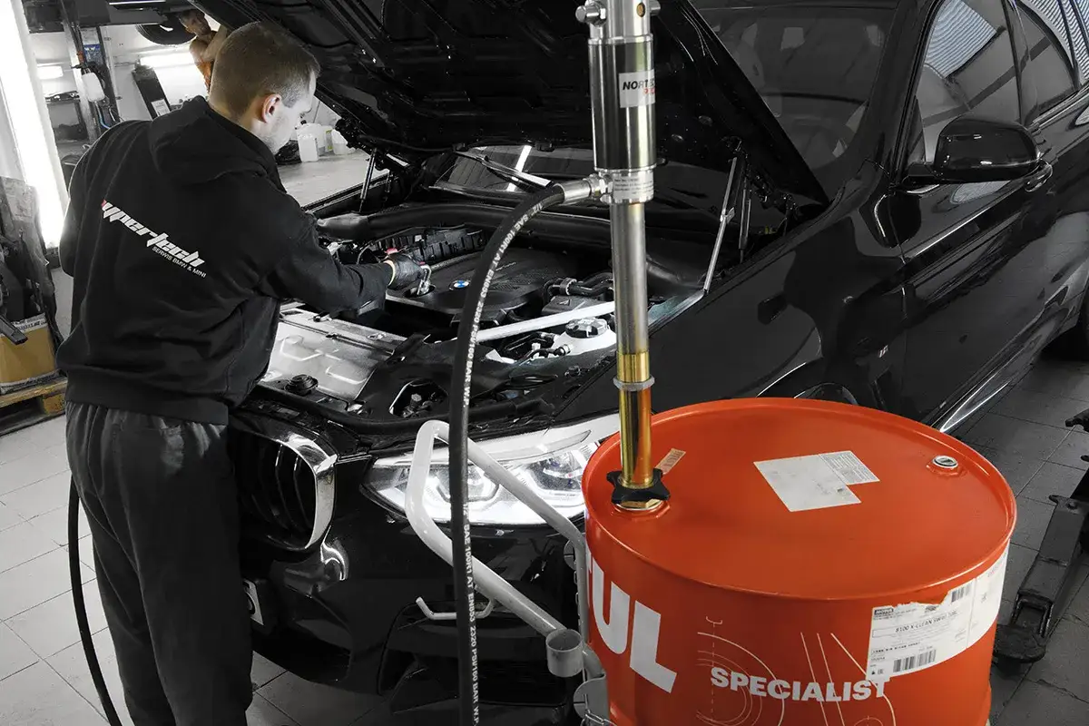 Mechanik wymienia olej w czarnym BMW. Obok stoi duży, pomarańczowy bęben z olejem. Dowiedz się, ile kosztuje wymiana oleju robocizna.