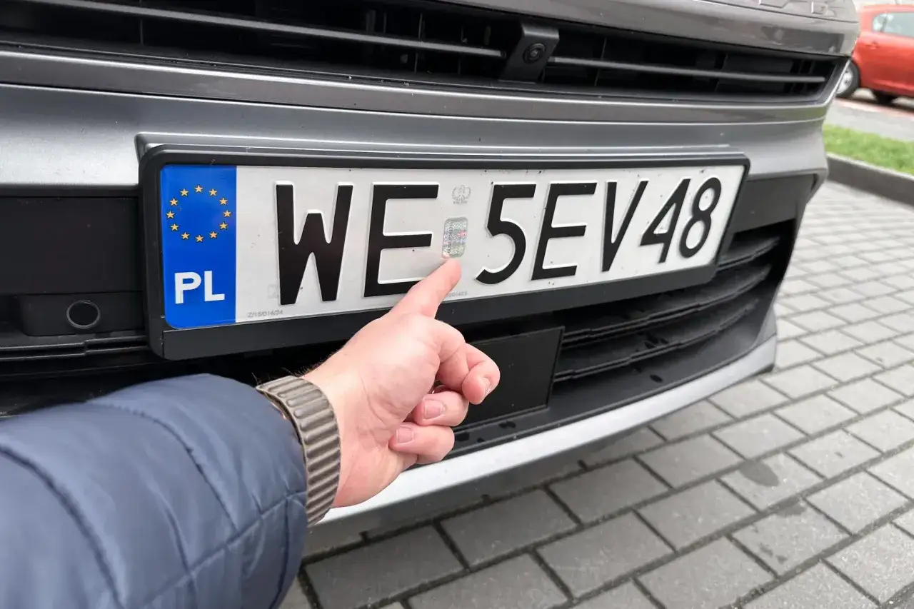 Dłoń wskazuje hologram na tablicy rejestracyjnej. Zastanawiasz się, ile kosztuje przerejestrowanie samochodu?