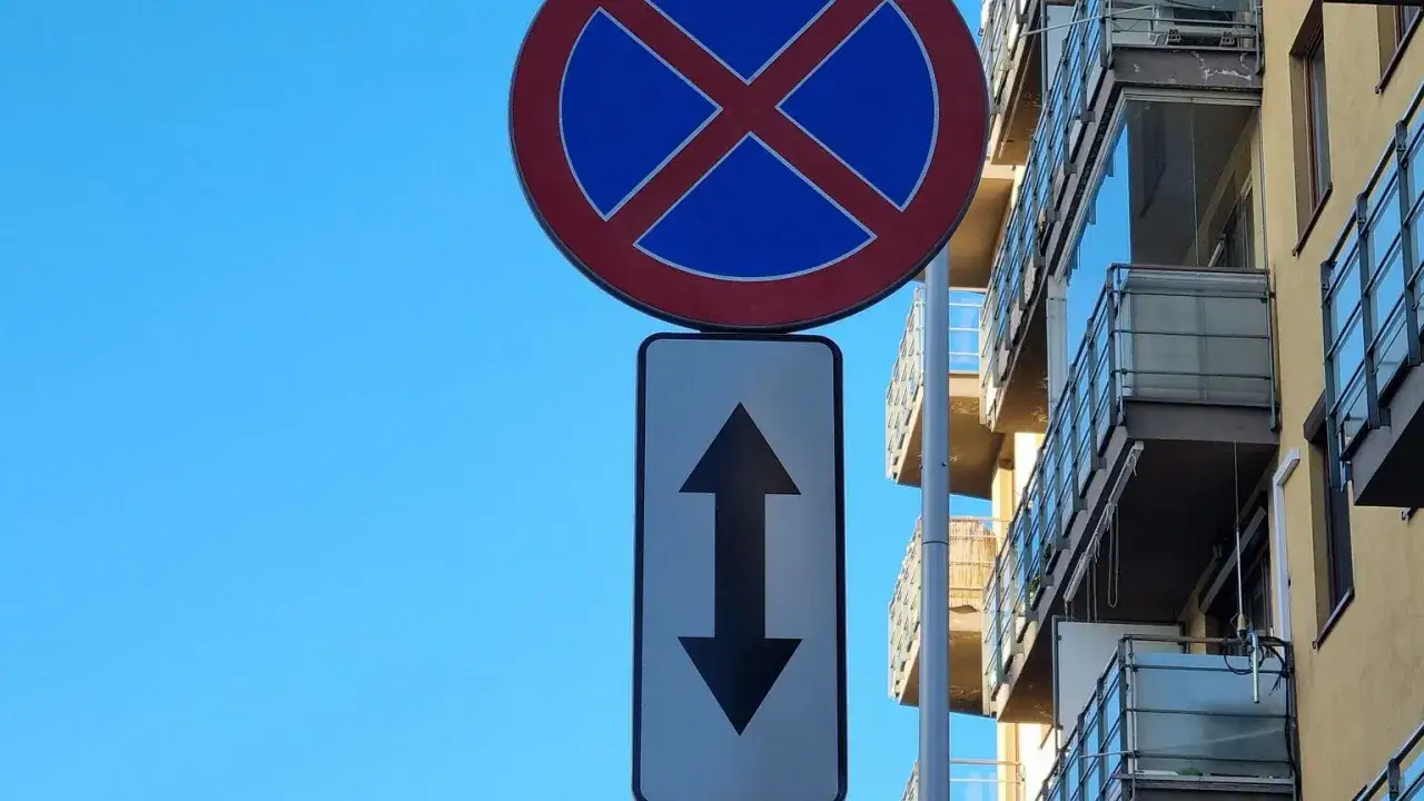 Znak zakaz zatrzymywania z tabliczką wskazującą zakaz w obu kierunkach. Obok nowoczesny budynek z balkonami.